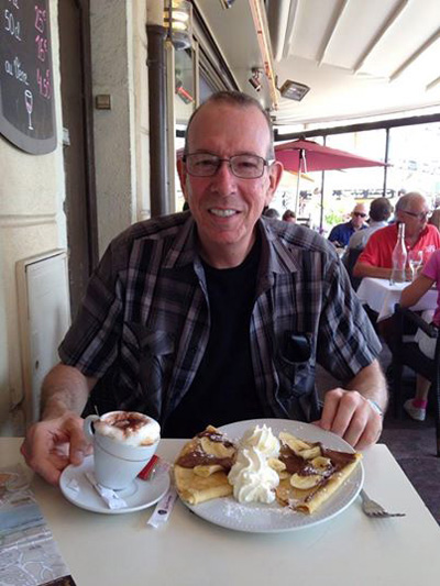Photo shows Gene smiling with a plate of crepes covered with whipped cream, and a mug of coffee also covered with whipped cream.